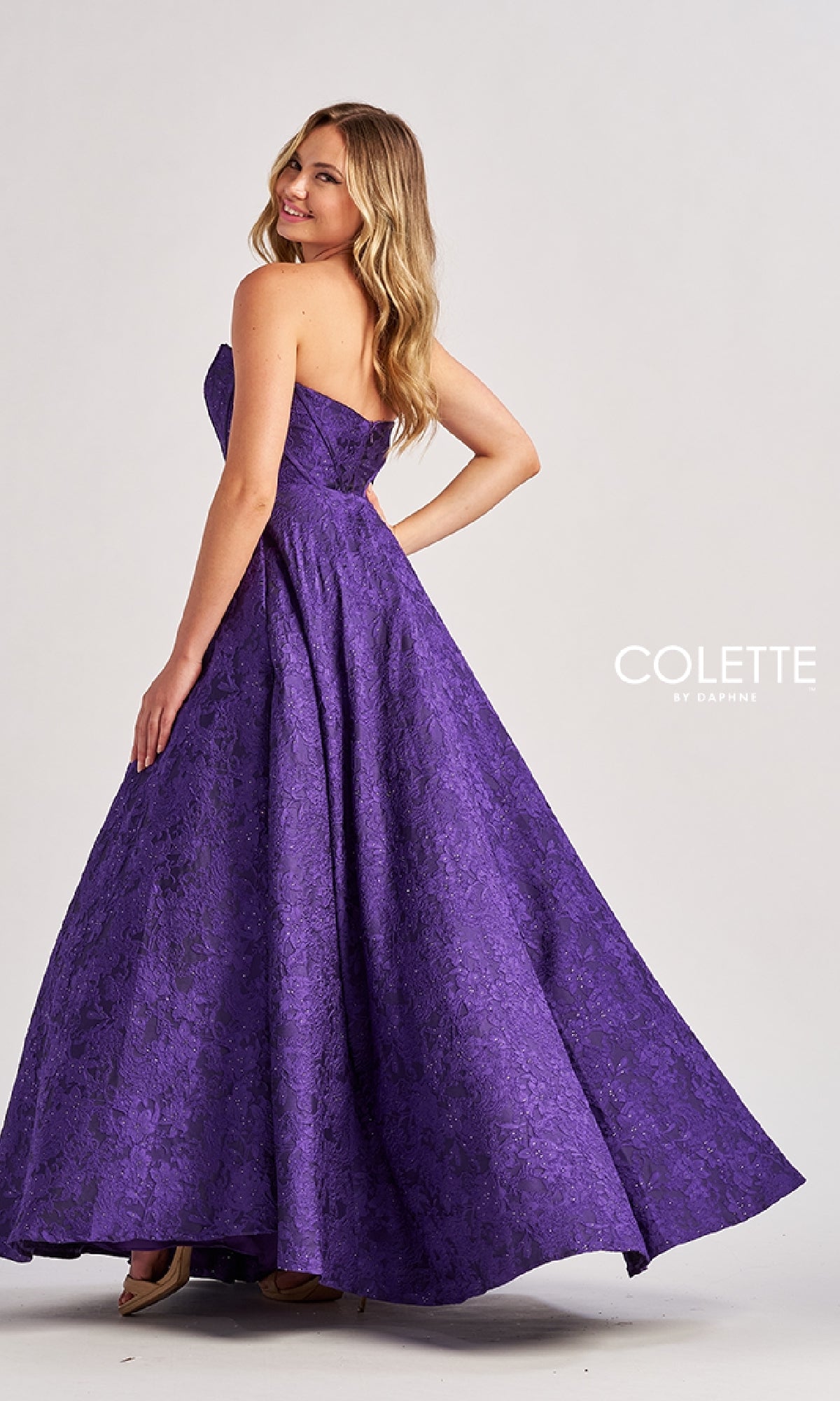 A woman poses in the floor-length purple Colette CL8675 prom dress with an A-line skirt, textured pattern, and pockets, standing against a plain light background.